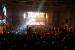 photos taken from the back of the Strand Theatre balcont of a sold out crowd. One man is standing with his hand in the air.