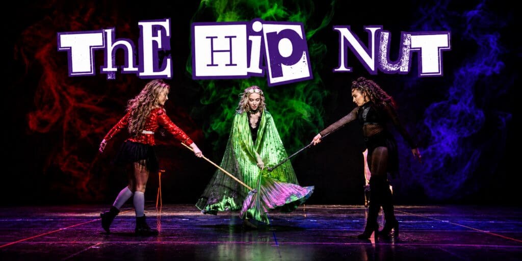 Three performers in colorful costumes sweep a stage with brooms beneath a large “The HIP NUT” title, surrounded by red, green, and blue theatrical smoke.