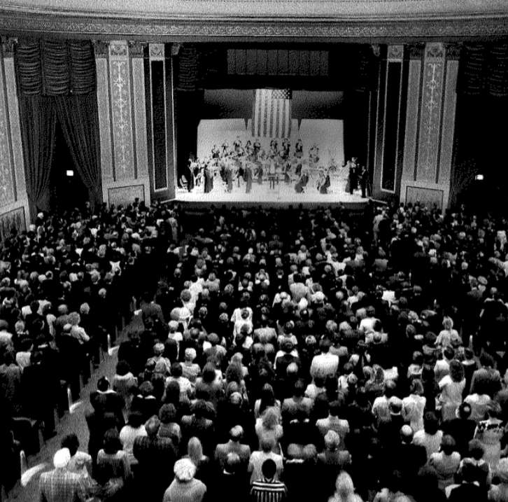 A black and white photo of the interior of the Strand Theatre during its 1980 Reopening Gala with Ella Fitzgerald and the York Symphony Orchestra on stage.