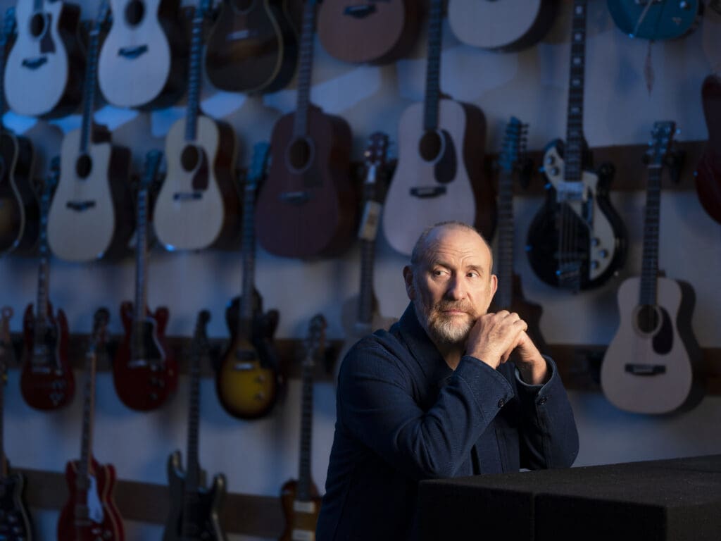 Colin Hay sitting with folded hands in front of an array of guitars