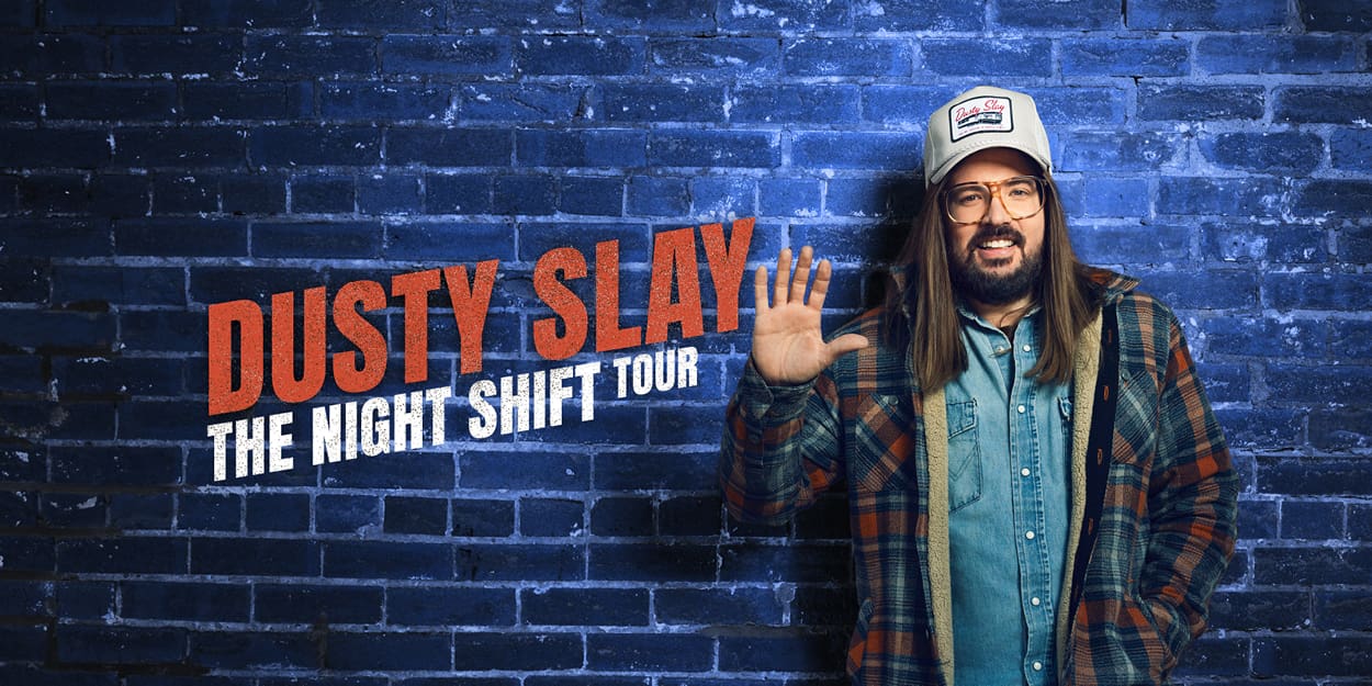 Comedian Dusty Slay standing in front of a brick wall waving with his name and tour title on the left.