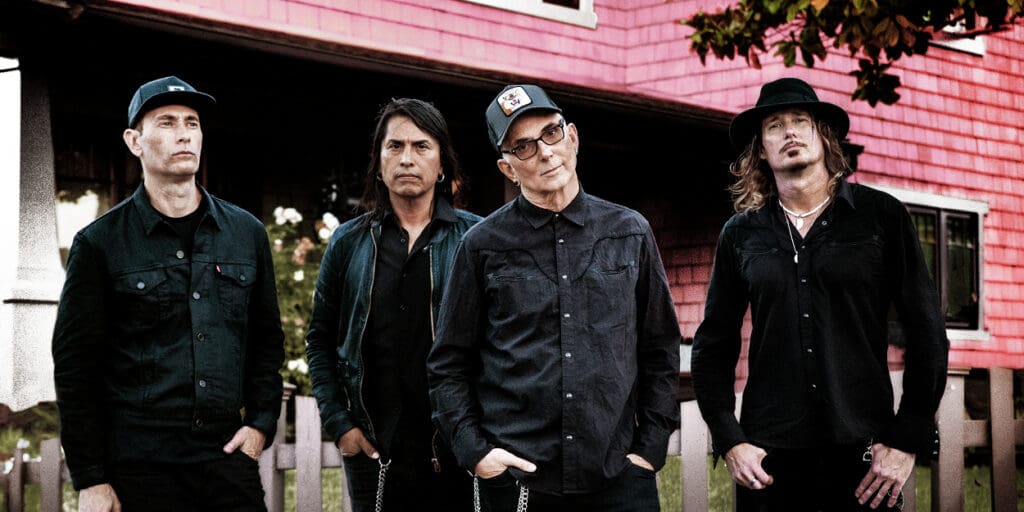the band Everclear standing side by side, all wearing black, in front of a red building