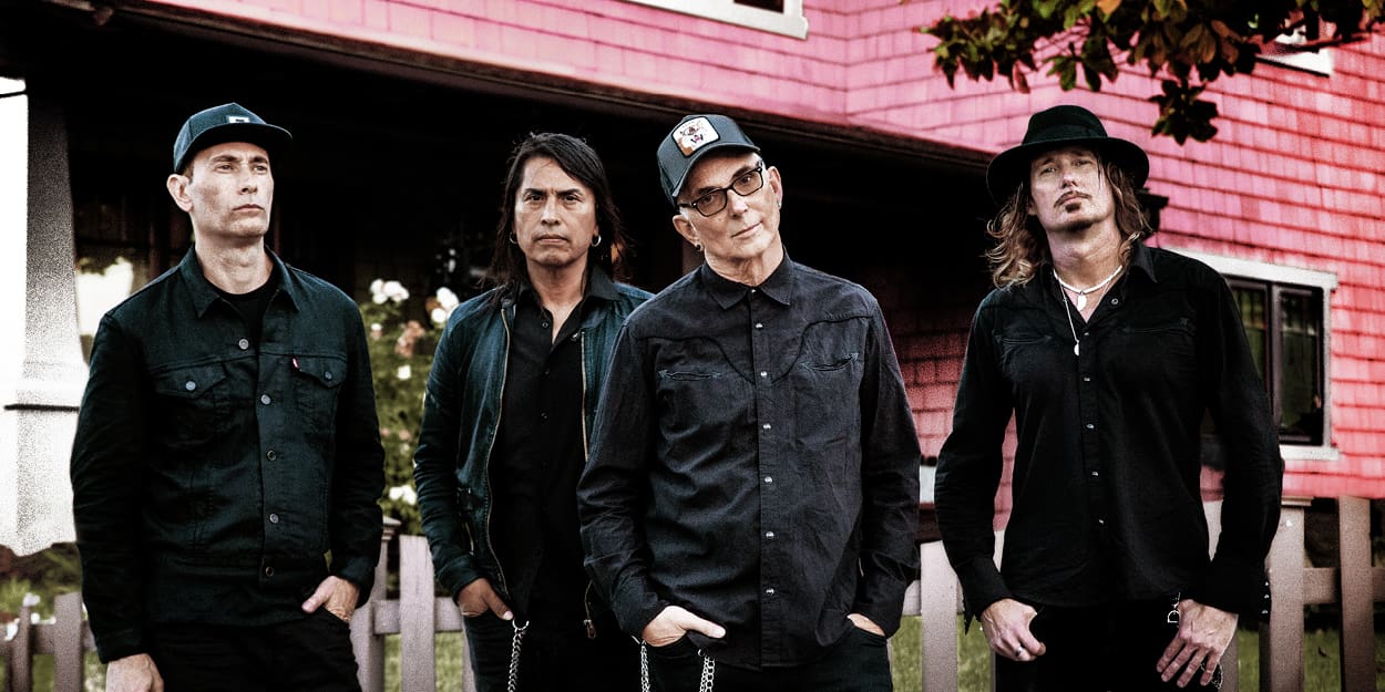 the band Everclear standing side by side, all wearing black, in front of a red building