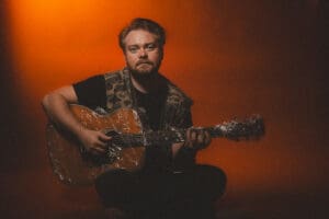 Singer/songwriter Guthrie Brown playing guitar against orange background