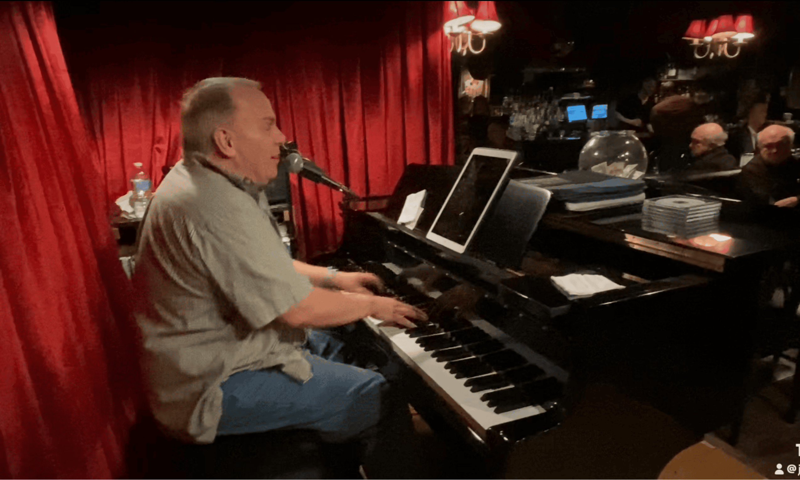 Joe Regan playing a baby grand piano on the left in front of red curtains with patrons listening on the right
