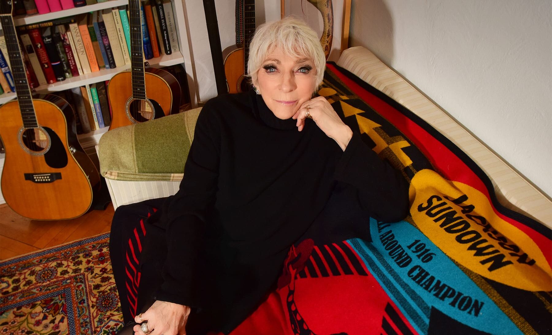 Judy Collins in black sitting on a couch with guitars behind her