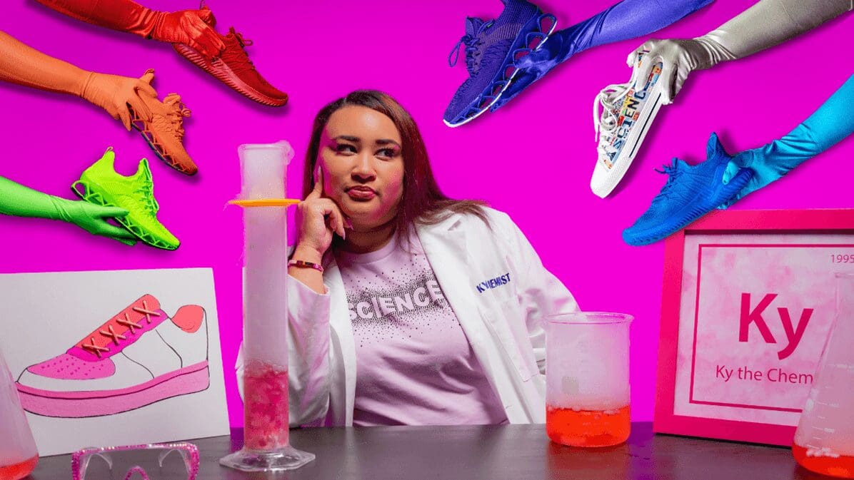 Ky the Chemist sits at a lab table with a hot pink background with colorf sneakers being held out around her