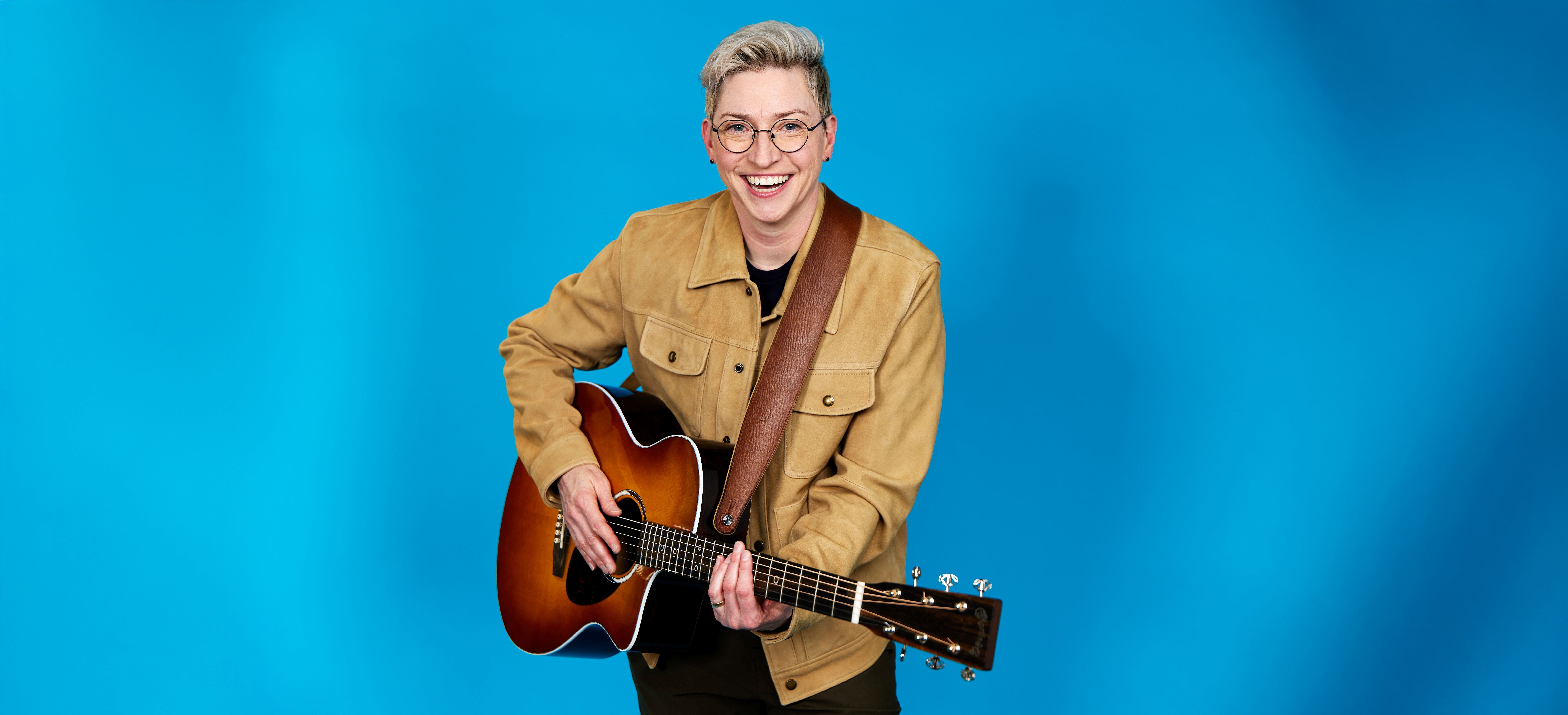 Comedian Kristin Key wearing a tan shirt holding an acoustic guitar against a sky blue background