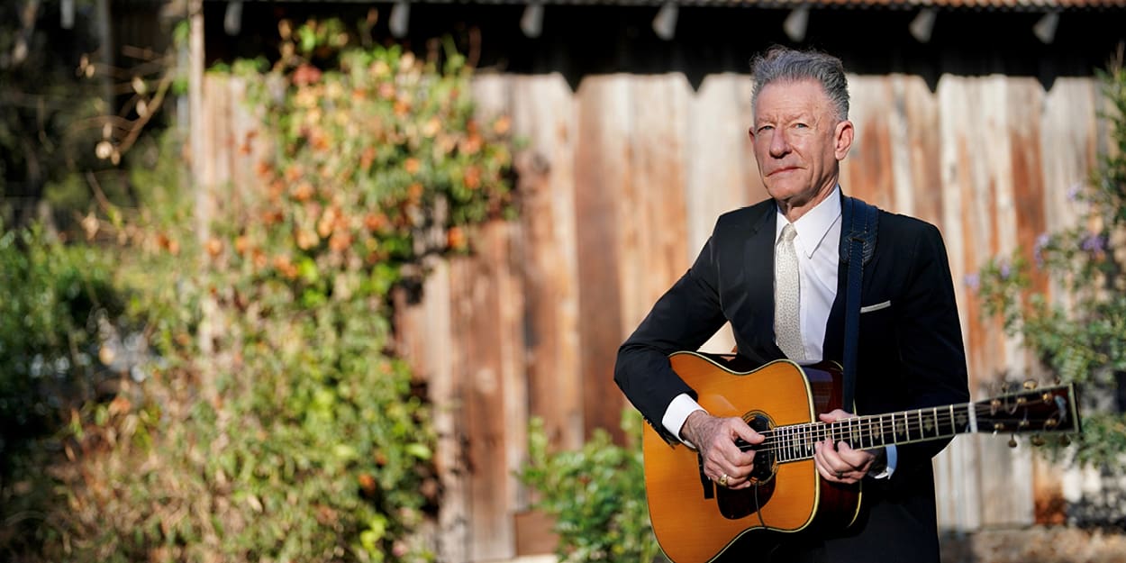 Lyle Lovett playing an acoustic guitar near a bush and brown wooden fence