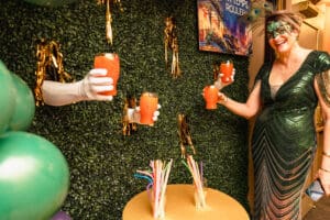 DIrector of Development and GALAvant wearing green and a painted mask poses infront of the drink wall at GALAvant: Mardi Gras in 2024.