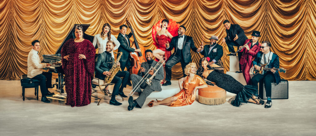 All members of PostModern Jukebox dressed in formal attire posing across a stage