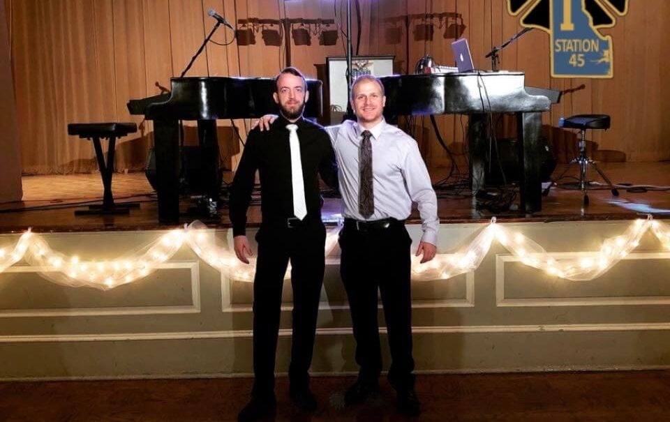 2 pianists standing shoulder to shoulder, one in a black short and white tie and the other in a white shirt and black tie
