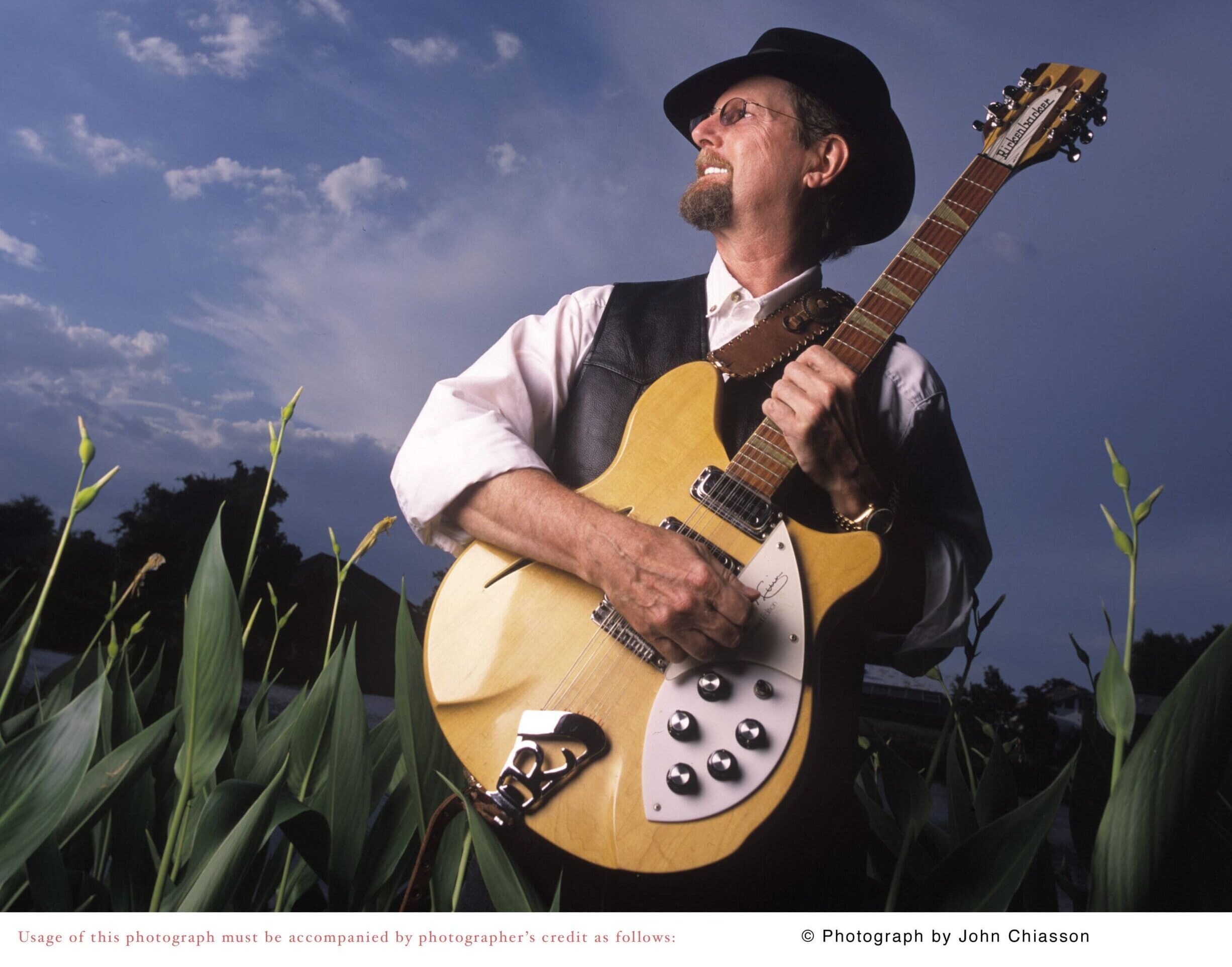 RogerMcGuinn holding guitar in a corn field with blue sky
