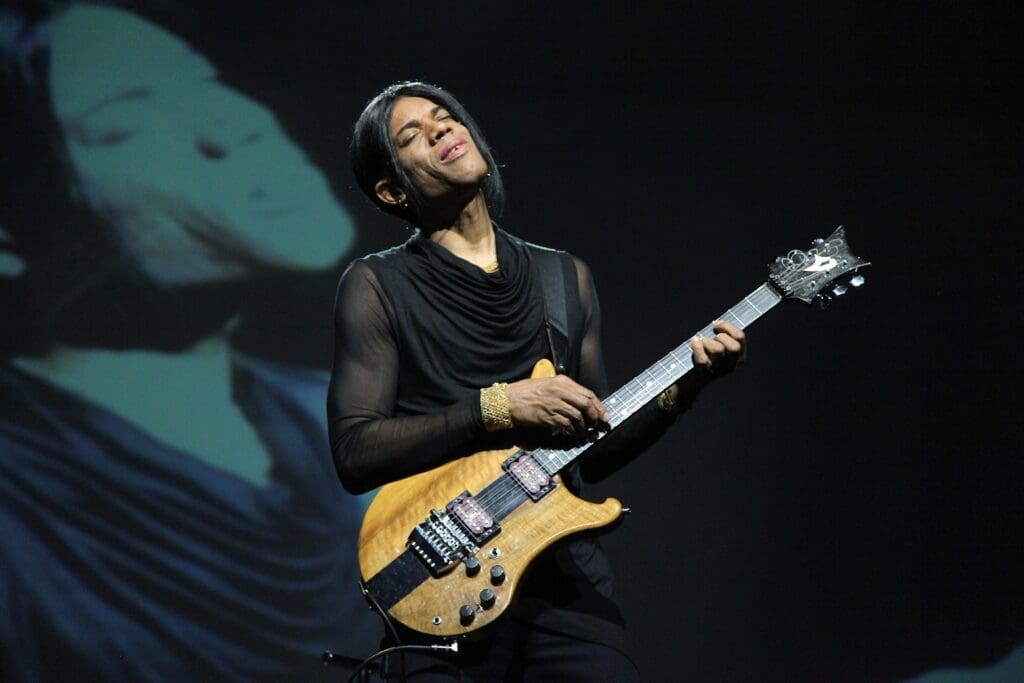 Stanley Jordan performing guitar on stage wearing all black with a projection of himself just behind on a screen.