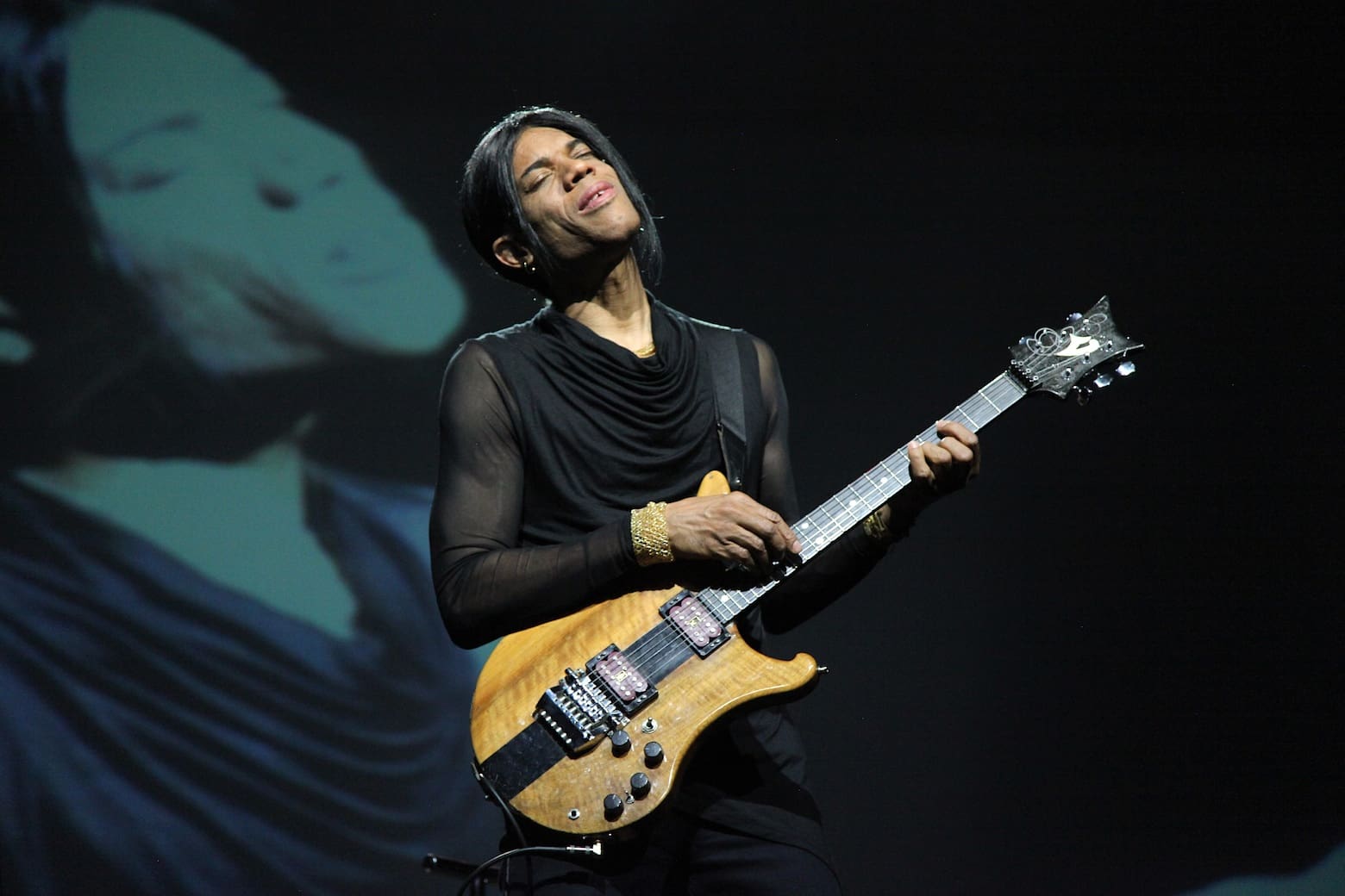 Stanley Jordan performing guitar on stage wearing all black with a projection of himself just behind on a screen.