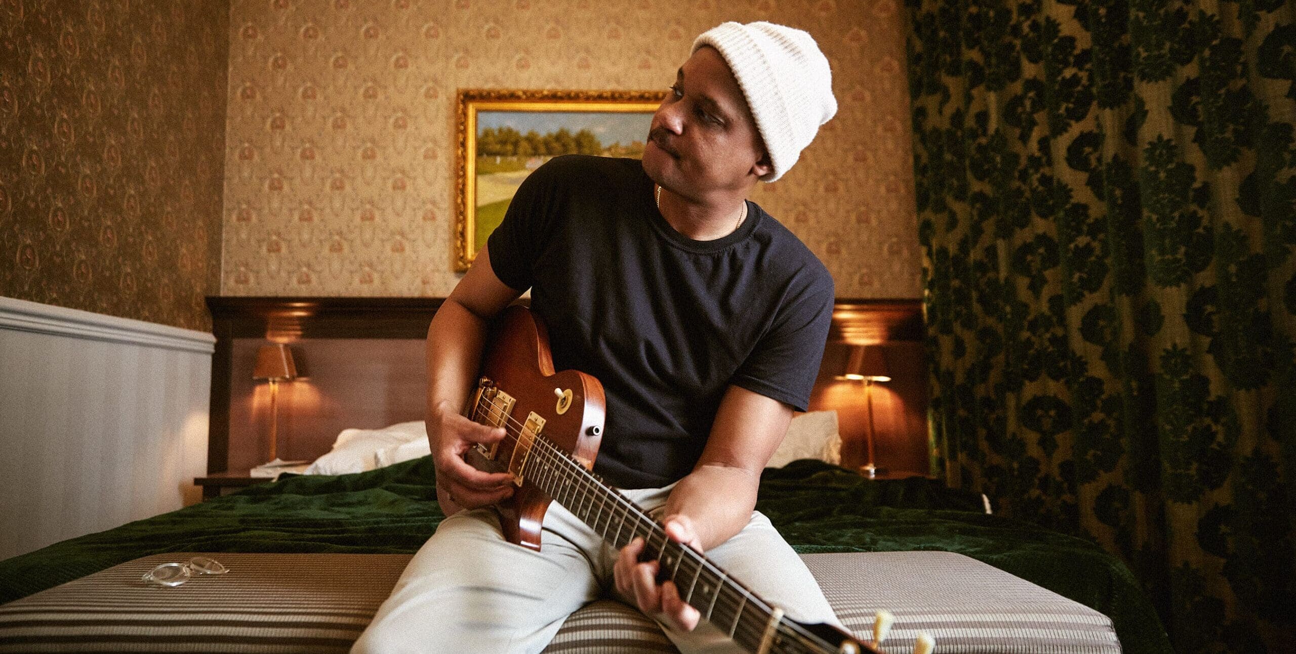 Son Little sitting on a bed holding his guitar and loooking to the left