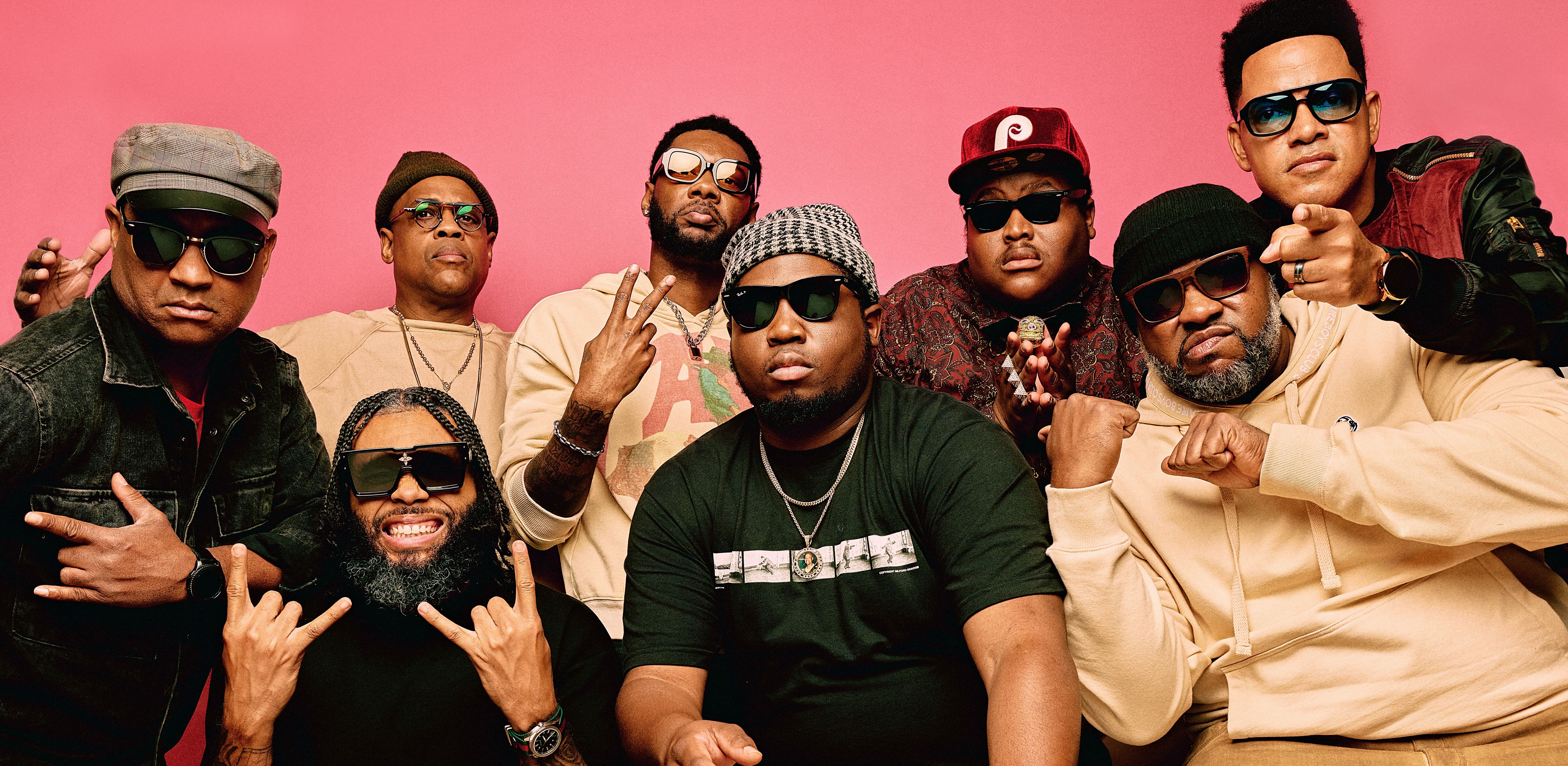 All 8 members of The Soul Rebels posing in front of a rose pink background