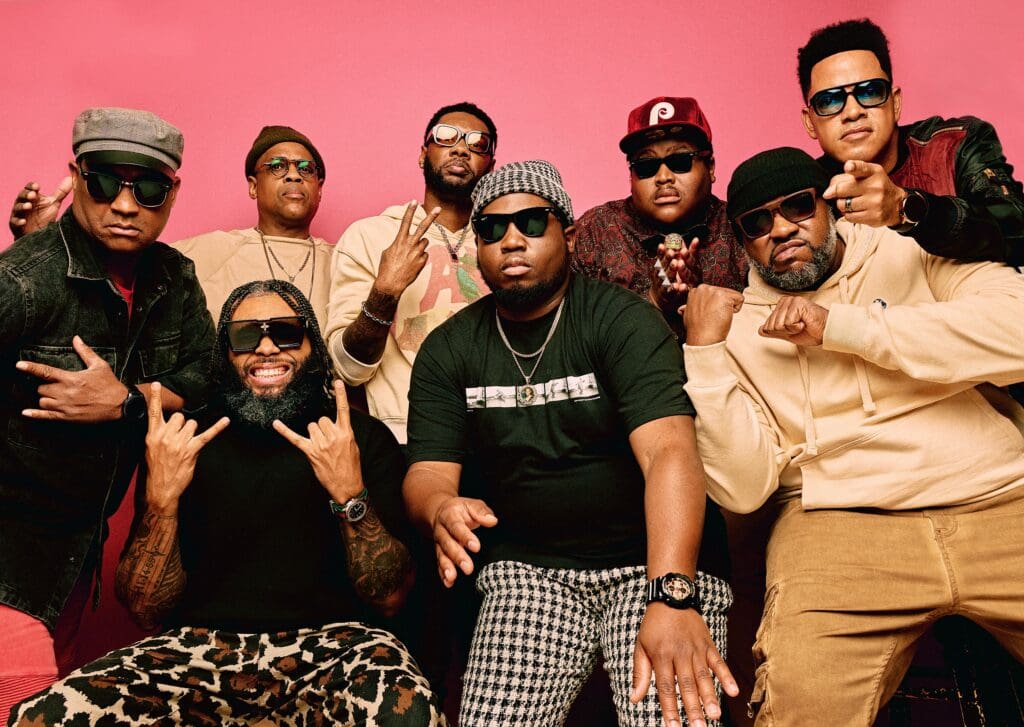 All 8 members of The Soul Rebels posing in front of a rose pink background