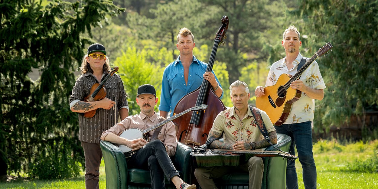Members of the band The Infamous Stringdusters holding their instruments in a green field with trees in the background