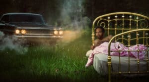 Tre Burt lying on a brass bed in a field wth car and headlights behind