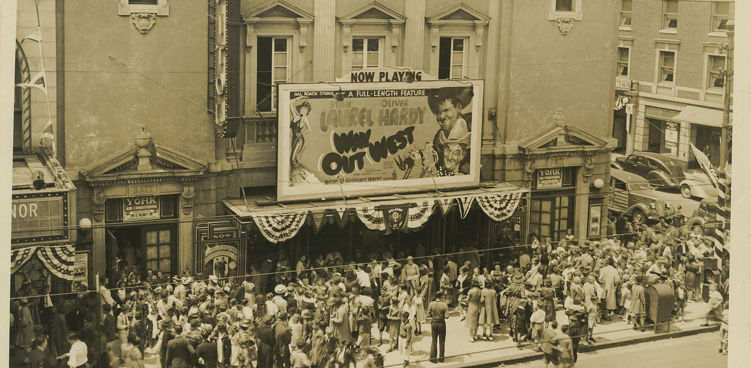An old, sepia toned image of the Capitol Theatre. Patrons stand in front of the theatre and look at the marquis where there is a "Now Playing" poster for the movie Way Out West featuring Lauren and Hardy.