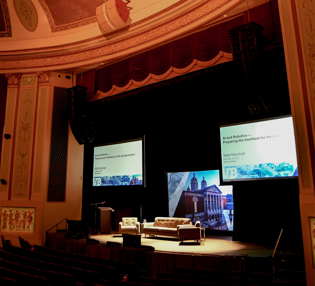 Image of the Strand Theatre stage set up for a presentation with digital screens, chairs, a sofa, and a podium.